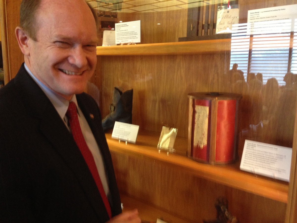 Sen. Chris Coons smiles next to one of the patent models he’ll feature in his office. It’s a red powder keg designed for DuPont in 1873.