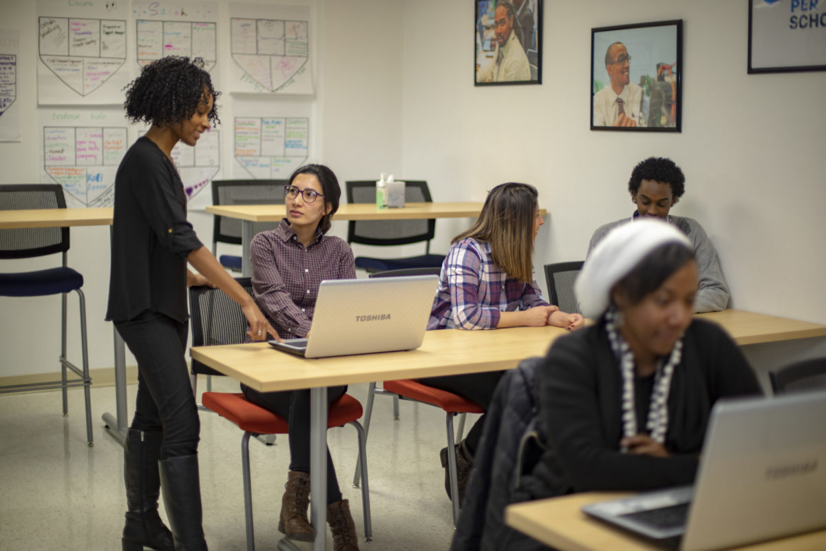 A student and teacher speaking at a tech training program for Per Scholas. Other students are at work around the classroom.