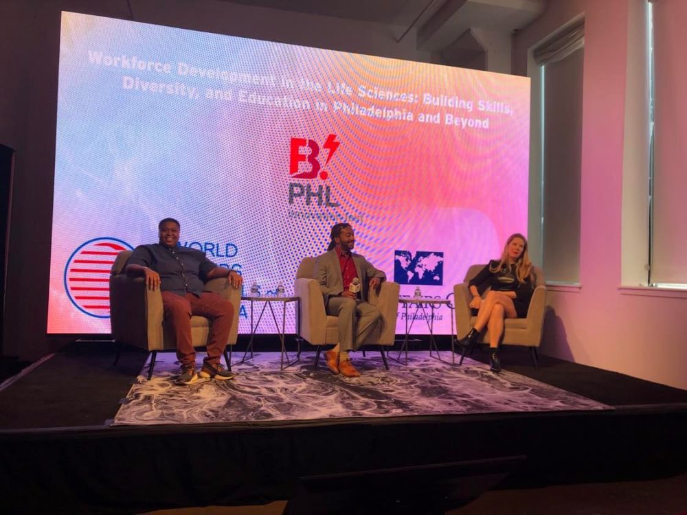 Phil Brooks, Tia Lyles-Williams and Lauren Swartz sit in chairs on the stage in front of a colorful background.