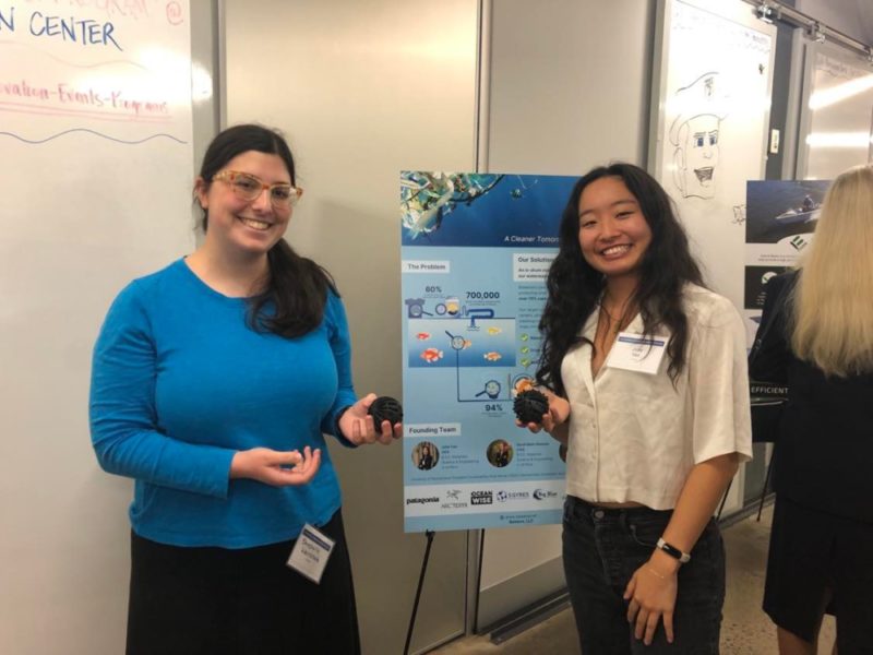 Shoshanna Weintraub and Emily Yan stand in front of a poster on an easel and holt their product