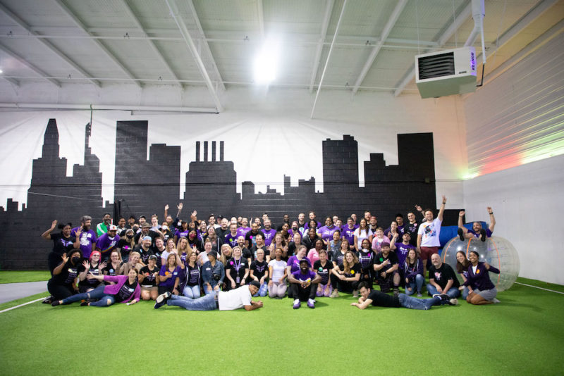 People in black and purple shirts on green turf in front of white wall with black Baltimore skyline.