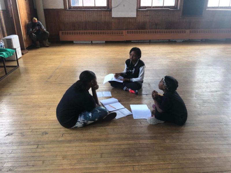 Three students sit in a circle on the floor with notebooks