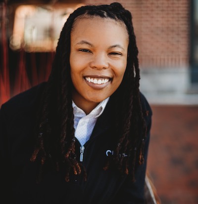 Woman with black dreadlocks smiles in black pullover and white shirt.