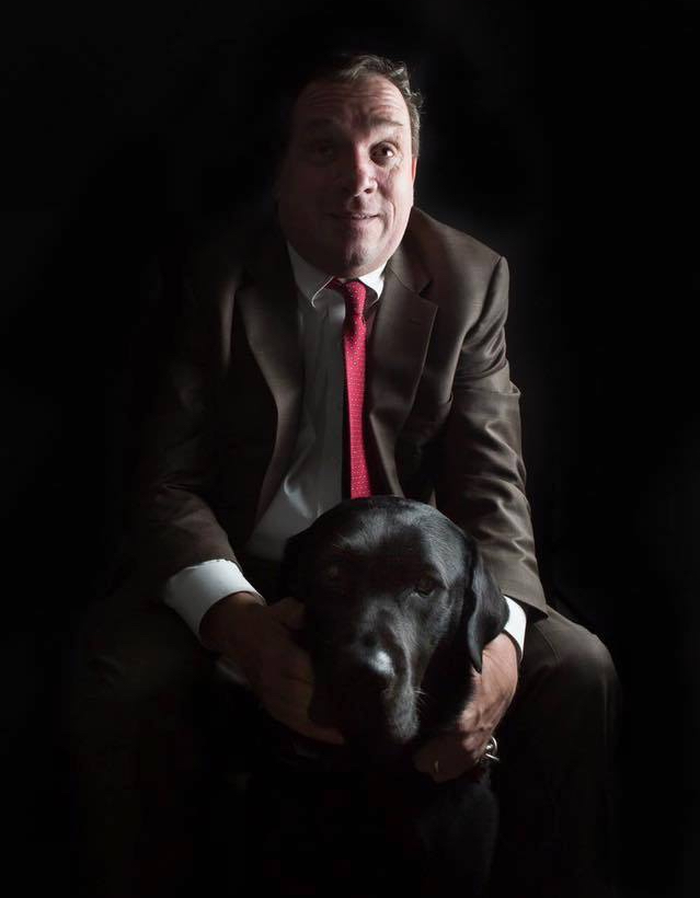 A man wearing a suit sits above a black labrador dog against a dark background.