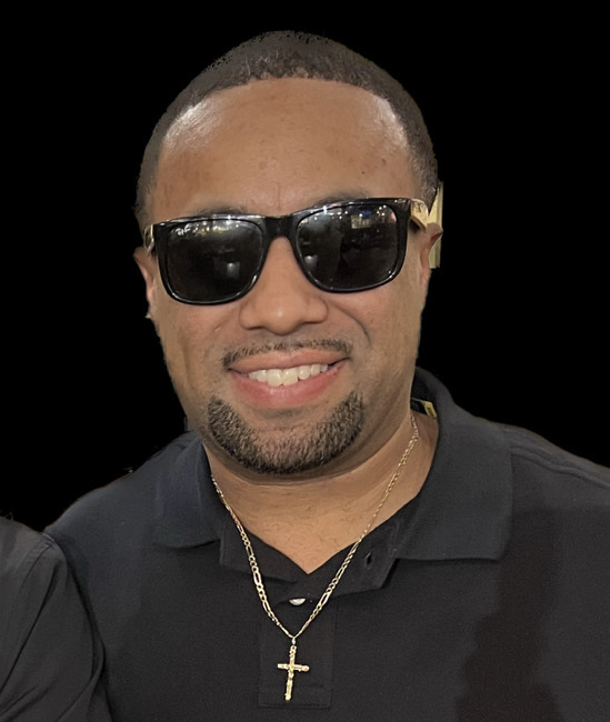 A man wearing dark sunglasses, a black shirt and a gold cross smiles.