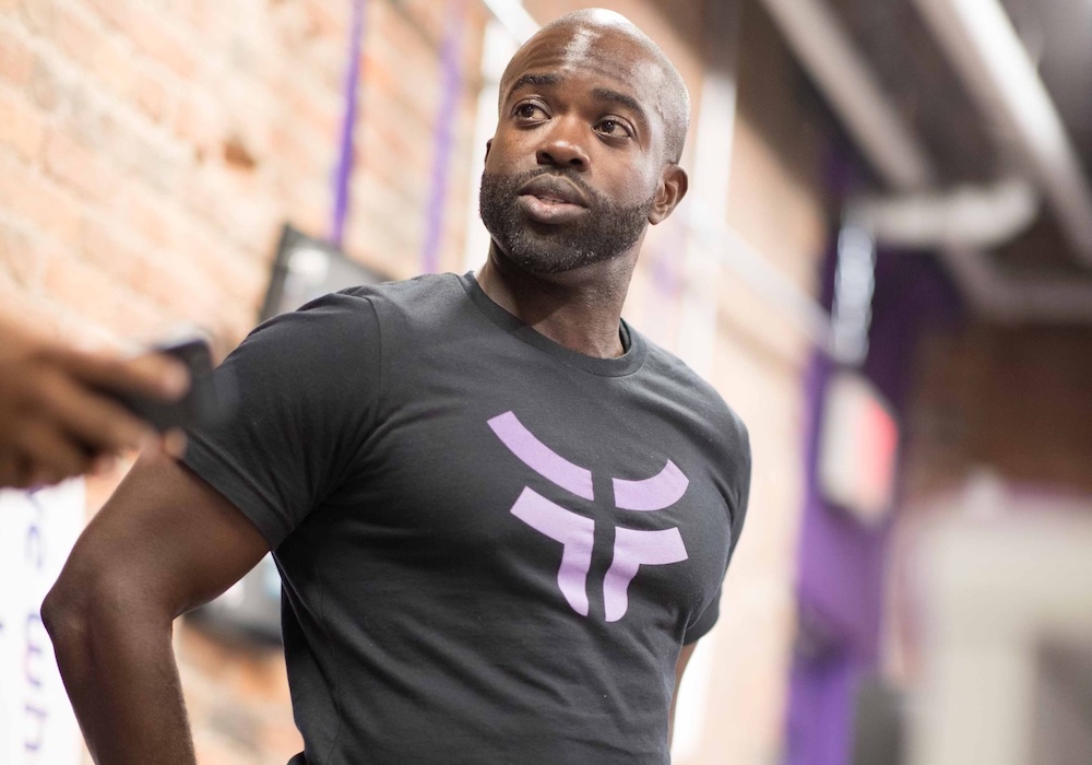 Man in black shirt with purple logo in front of red brick wall