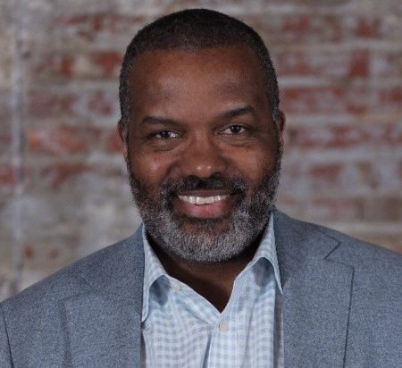 Man in grey suit smiles in front of red brick wall