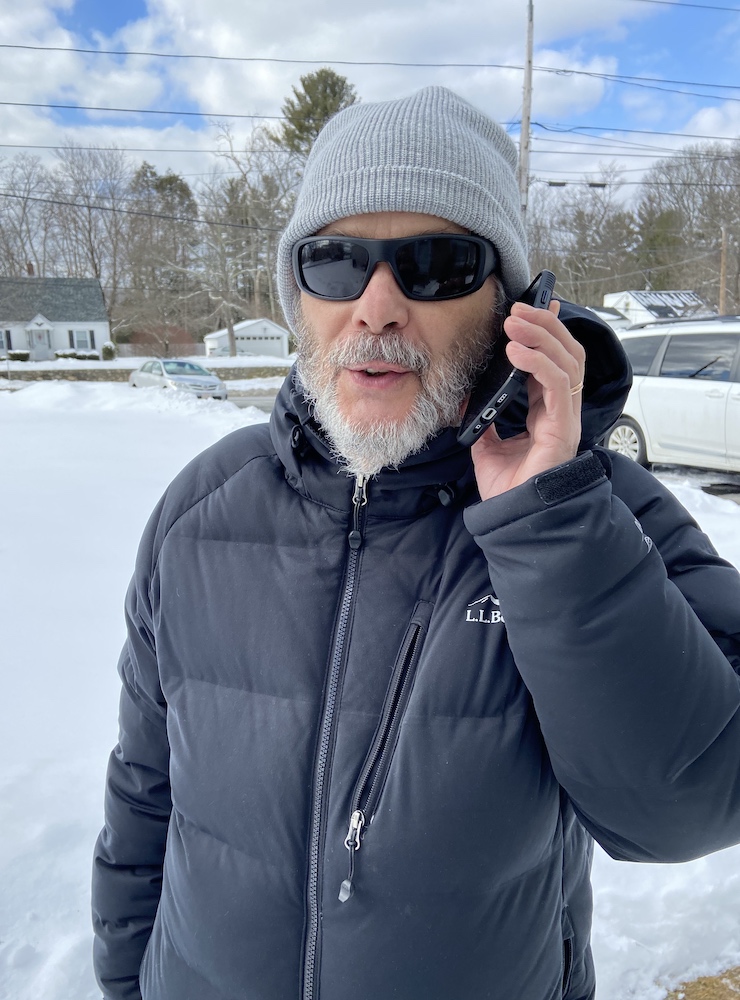 A man wearing a winter coat and hat and dark glasses holds a phone while standing in the snow.