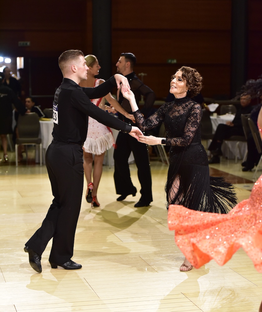 A man and woman in black formalwear dance in a ballroom.