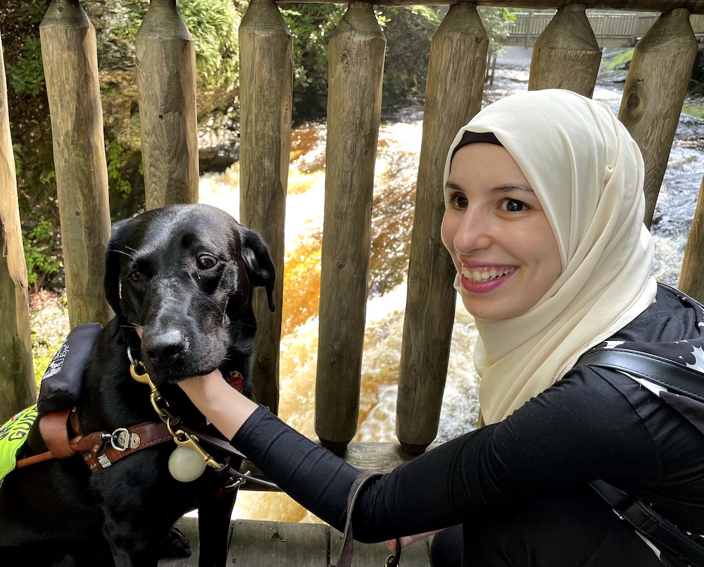 A smiling woman wearing a hijab and dark clothing kneels and holds a black service dog's head.
