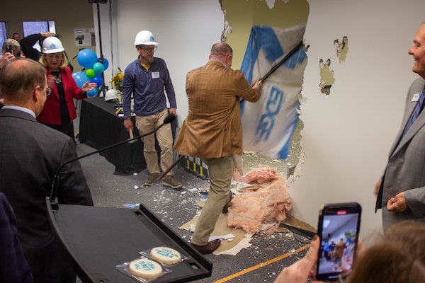 People stand as man in brown suit puts brown axe through beige and blue wall