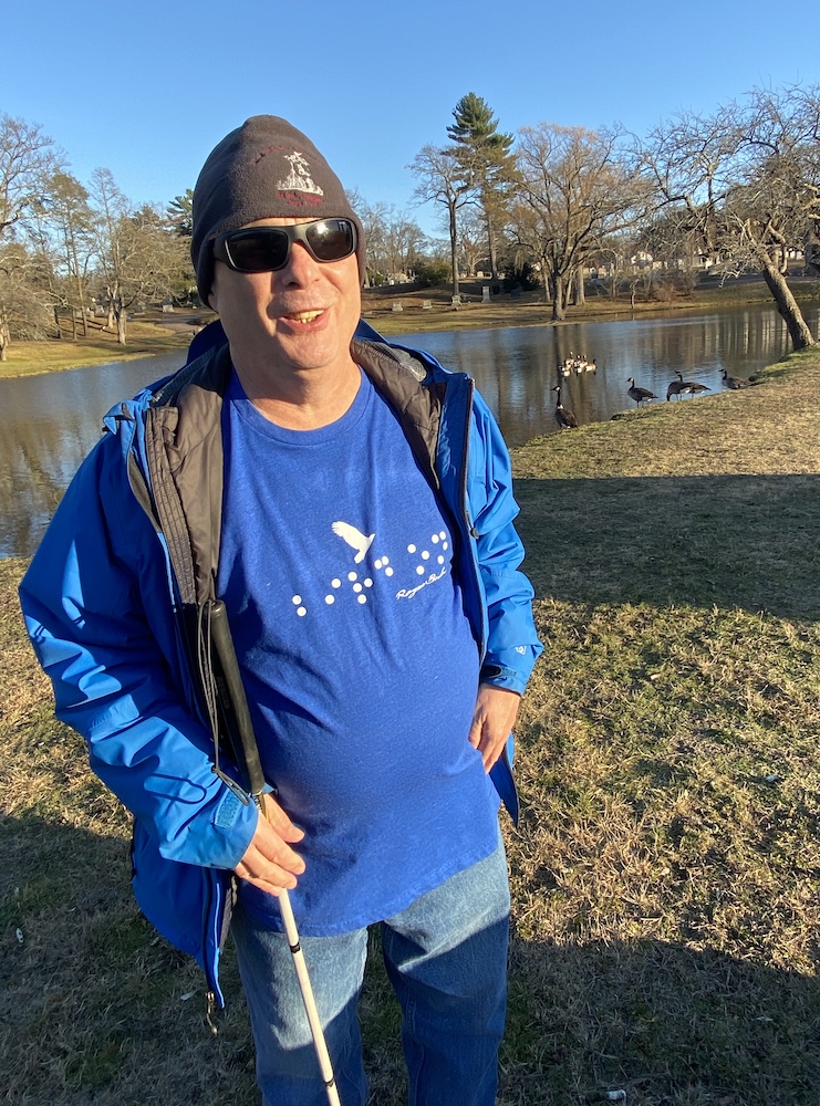 A man wearing a blue shirt, dark glasses and a hat stands with a cane beside a lake with geese in the background.