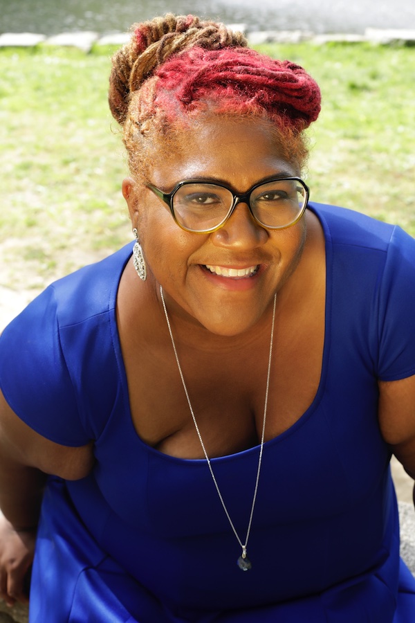 Woman with red and brown dreadlocks and glasses in blue dress in front of green grass