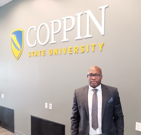 Man in grey suit poses by sign reading "COPPIN STATE UNIVERSITY" in white and yellow on grey wall
