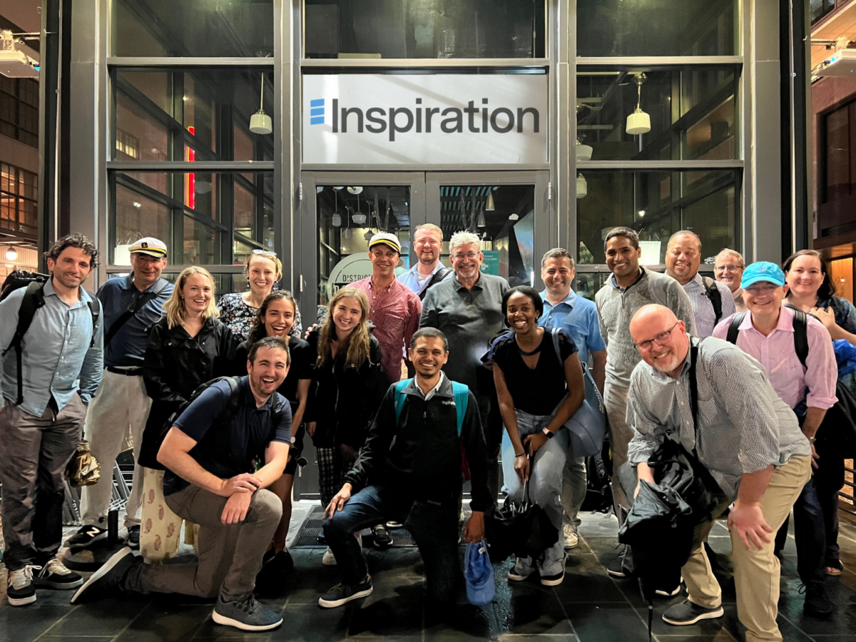 The Inspiration Mobility team poses in front of glass doors at a workplace summit. A sign featuring the company's name and logo hangs above. 