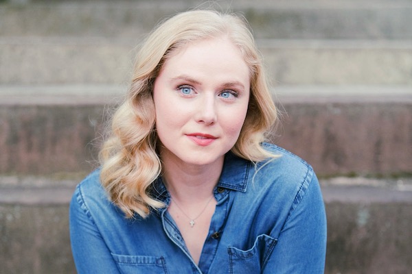 Woman with blonde hair in blue shirt in front of brown stairs.