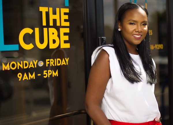 Woman with black hair in red blouse poses before glass with yellow text reading "THE CUBE"