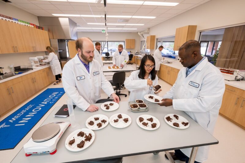 People in white lab coats at grey table with brown products on white plates in brown and white lab