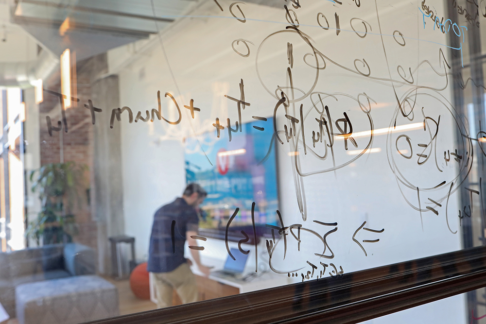 A man leaning over a computer to type while standing. A window with mathematical writing in the foreground.