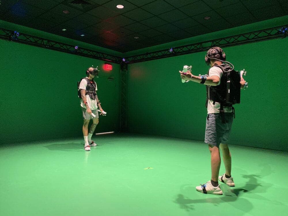 Players stand facing each other in a green room with VR headsets, motion sensors on their ankles and wrists and backpacks. 