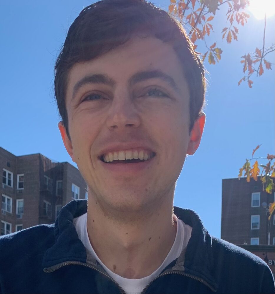 A headshot of Holden Foreman, who wears a blue zip-up and stands in front of a few apartment buildings.