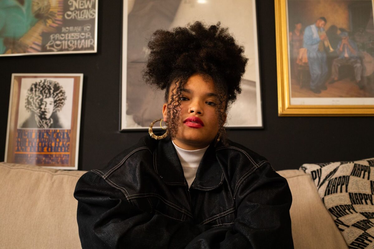 A young black woman sits on a couch in front of a black wall covered with posters and paintings. The woman has her hair tied up on top of her head. She is wearing a white turtleneck, a dark jean jacket, and gold hoop earrings.