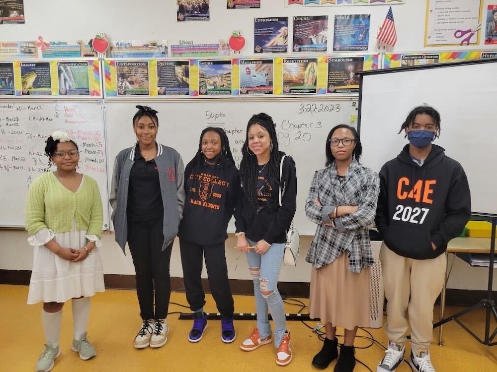 Children and teacher in multicolored clothing before whiteboard with black text on brown wood floor