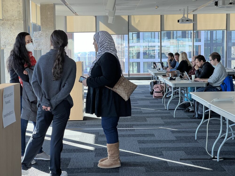 Three women stand in the front of an Amazon office space. Other technologists work on laptops at tables behind. 