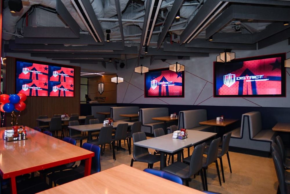 Tables and chairs in a restaurant-style setup. Screens with the District E logo are on the wall