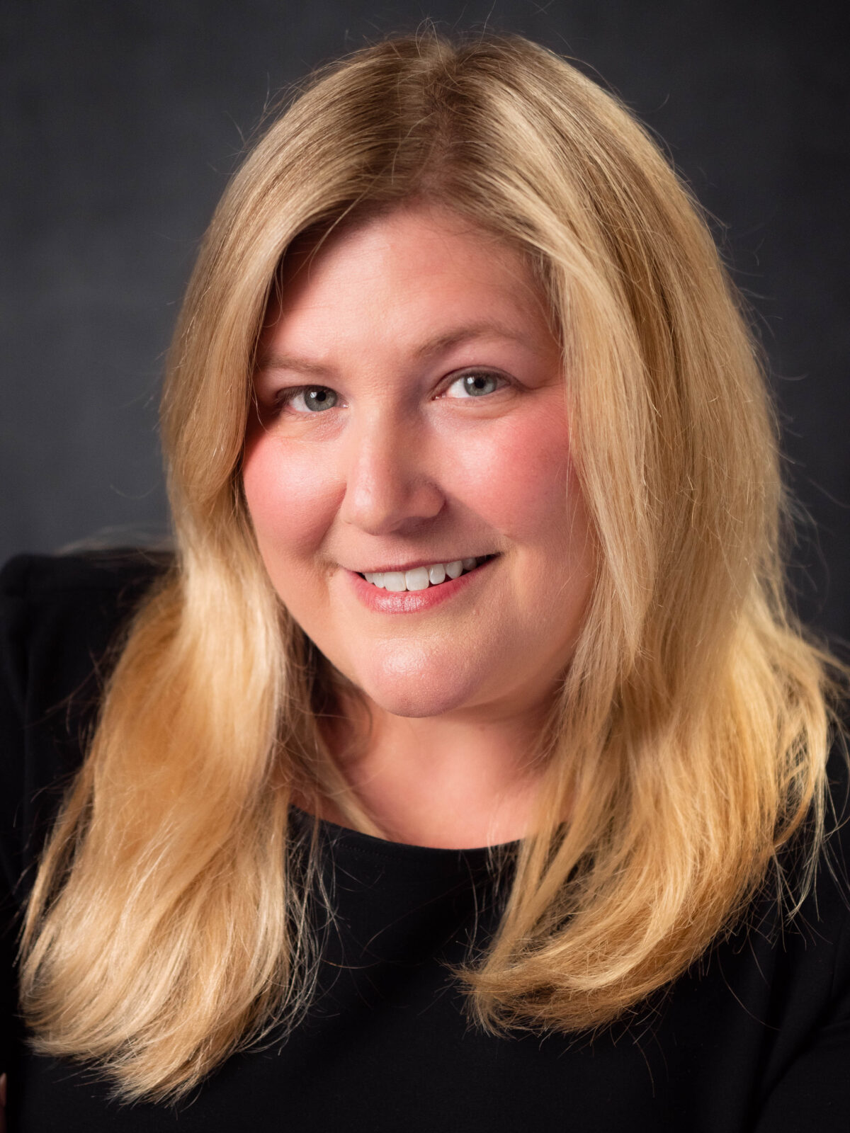 Beth Blauer with blonde hair and black shirt in front of black background.