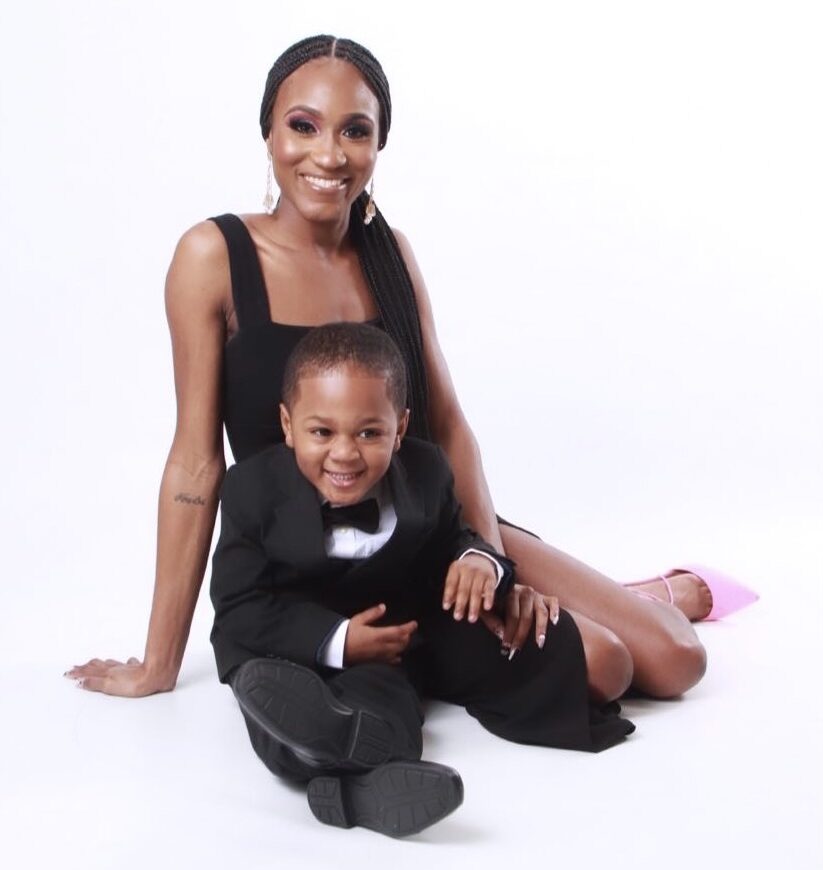 DeMia sits on the floor in front of a white background with her son. Both are in fancy black dress.