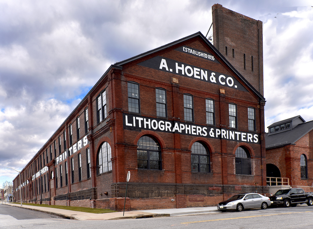 Brown factory building with white text on black background near blue sky with grey clouds.