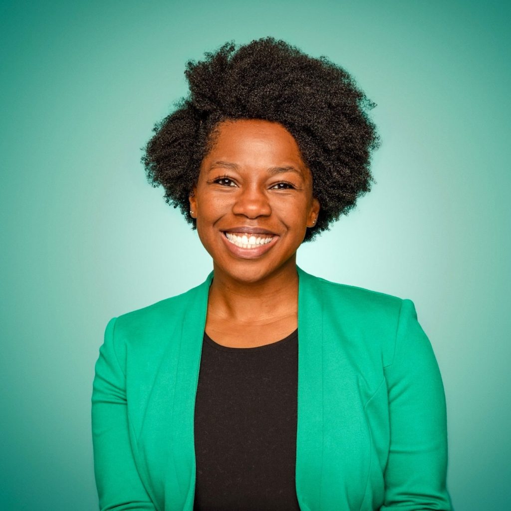 A headshot of Nadia Anderson, who wears a green blazer and stands in front of a green background.