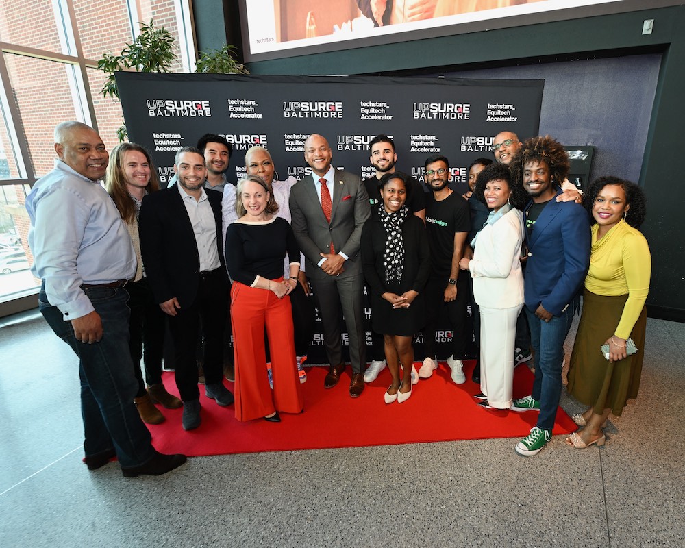 People in multicolored clothes stand on red carpet before black sign with white text