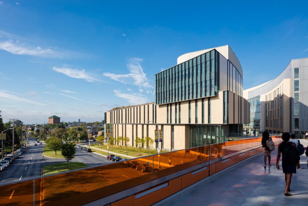 Beige academic building with turquoise windows in front of blue sky with white clouds near grey walkway with people.