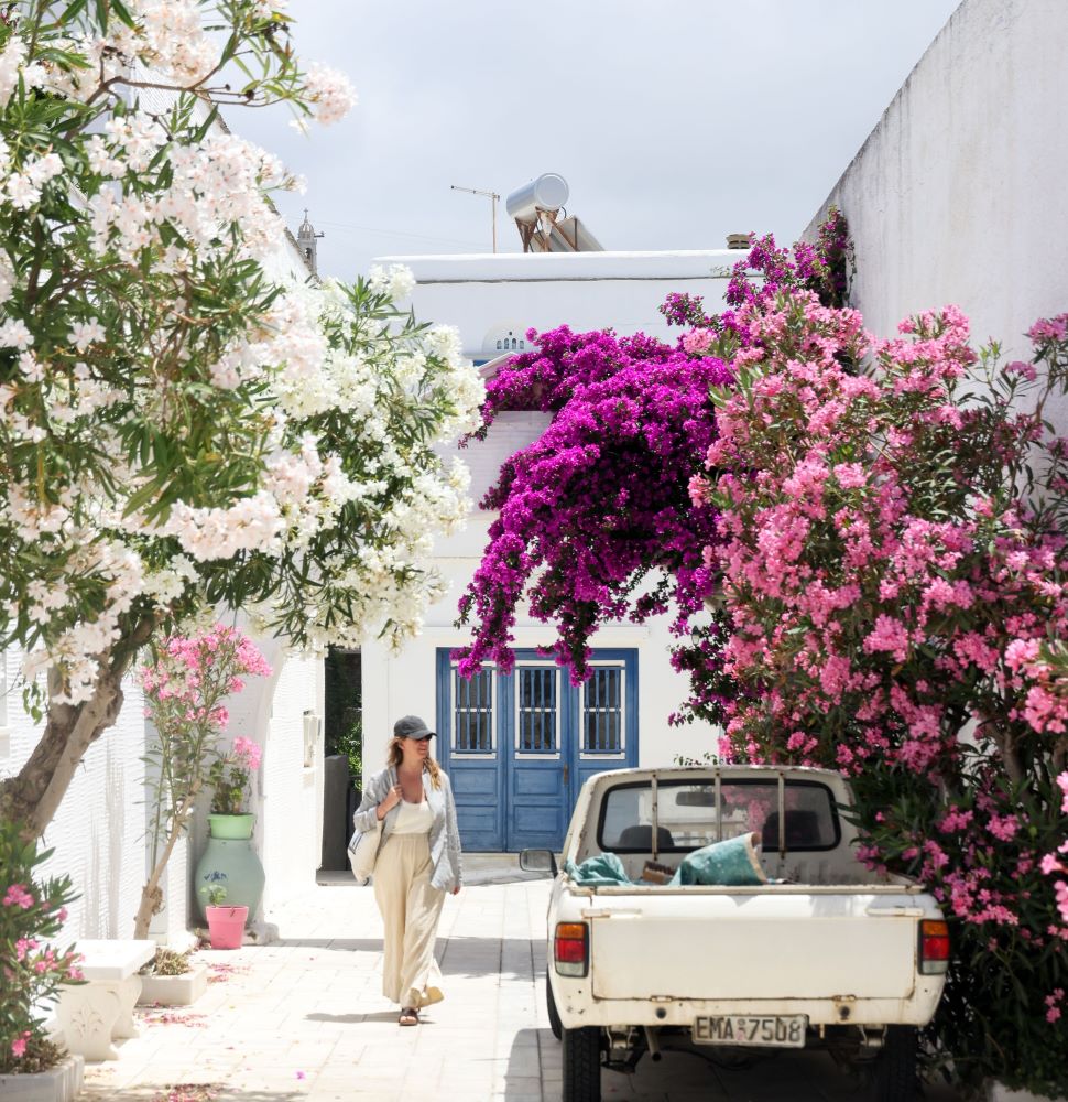 Katalina Mayorga stands in a driveway in Greece next to a white pickup truck. Blooming pink trees are on either side and a blue doorway is in the background.