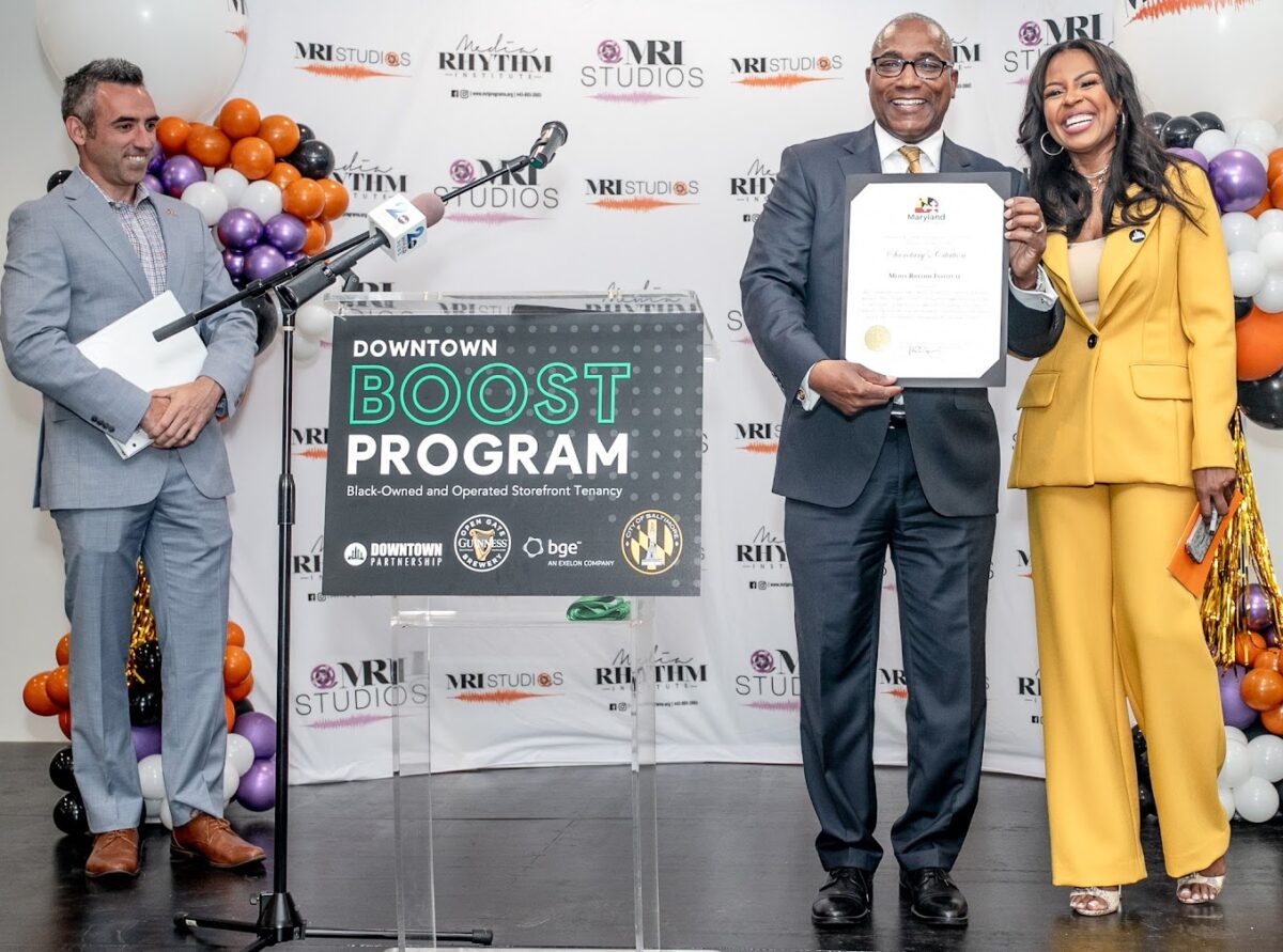 Shelonda Stokes in yellow suit with honorific certificate held by man in charcoal suit near man in grey suit