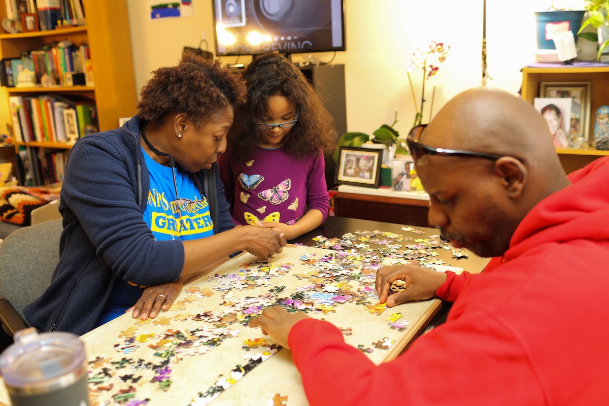An adult woman and man put together a puzzle while a young girl stands beside them.