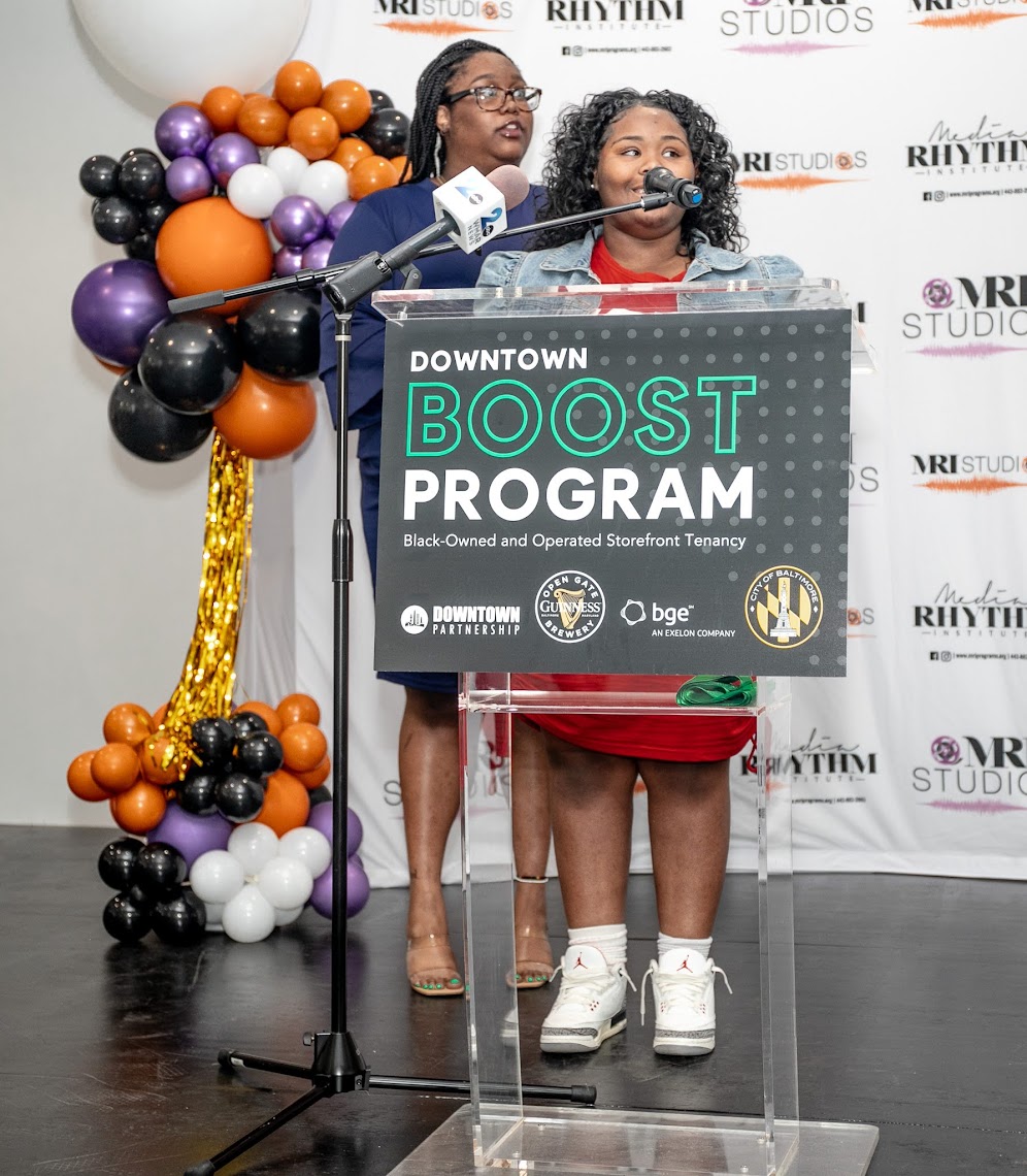 Ms. Kae stands behind Ms. Brooks, who speaks at a podium