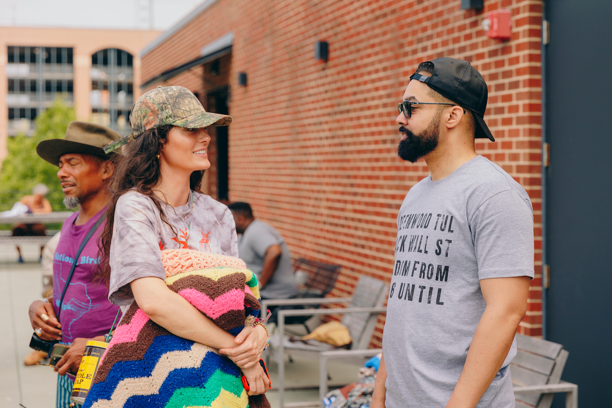Jasiri X in grey shirt with black text and black backwards hat speaking to woman in multicolored clothing and green hat