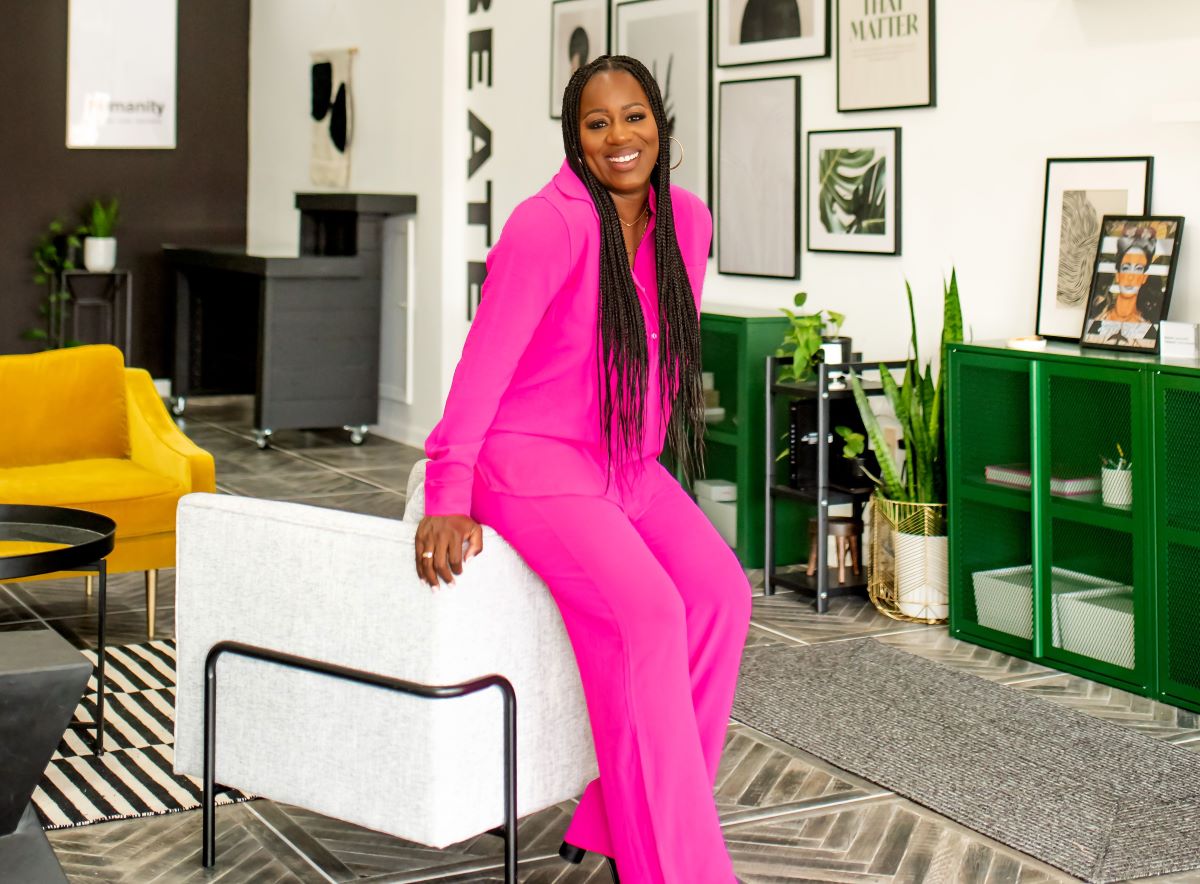 Jasmine Smith stands next to a chair in the Open Door Concept space, wearing a pink suit. 