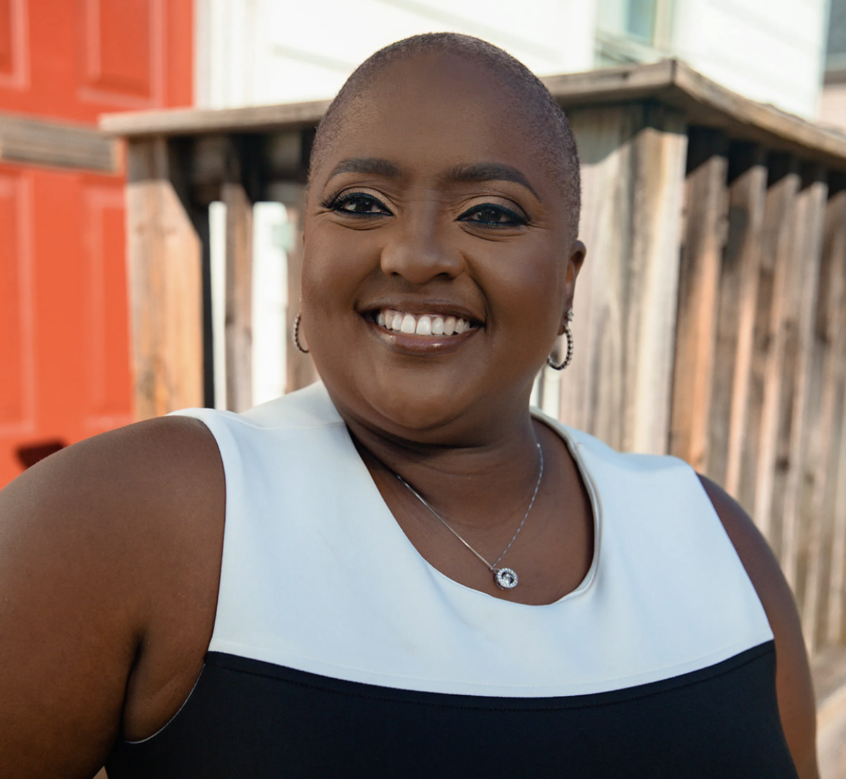 A Black woman stands in front of a wooden structure wearing a black and white top.