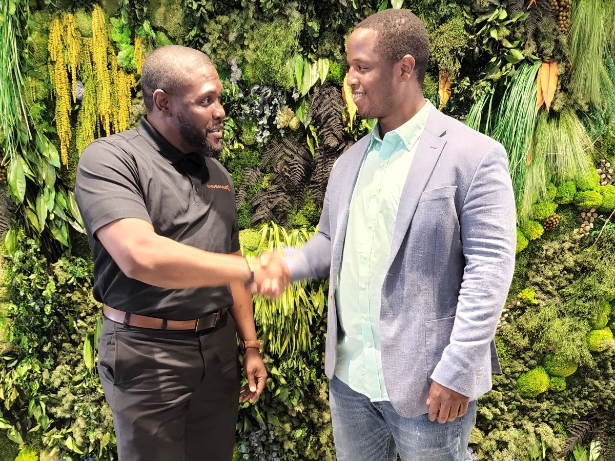 Two men in suits stand before a plant-filled background, shaking hands. 