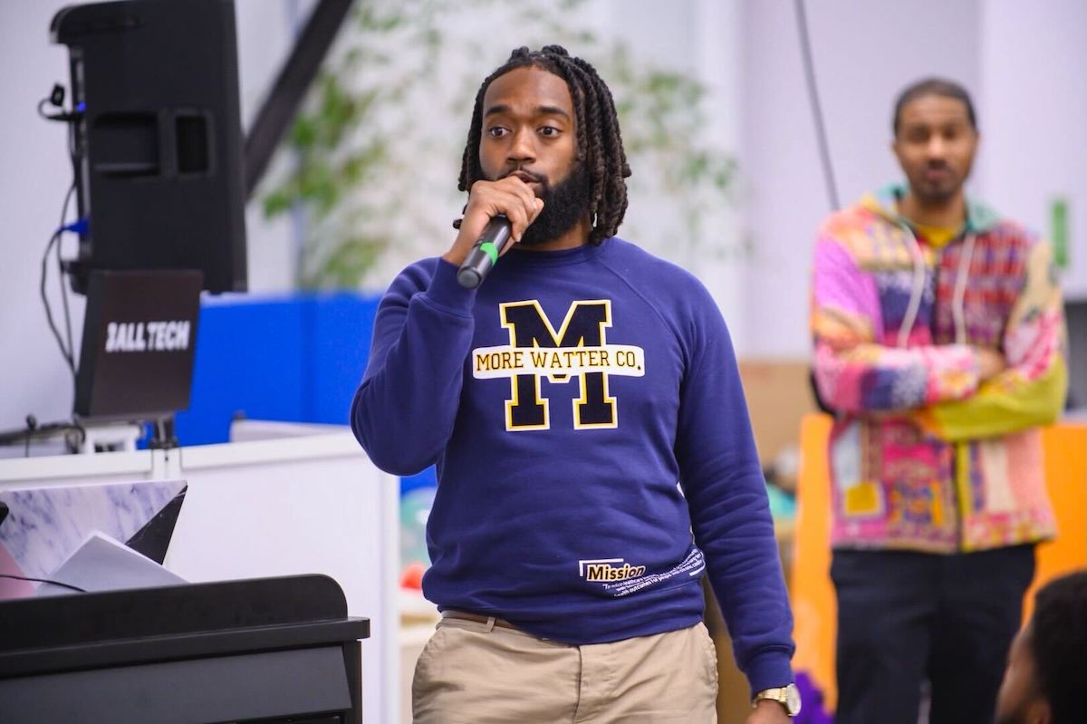 Anthony Watters holds microphone while wearing purple sweatshirt reading "MORE WATTER CO." in yellow