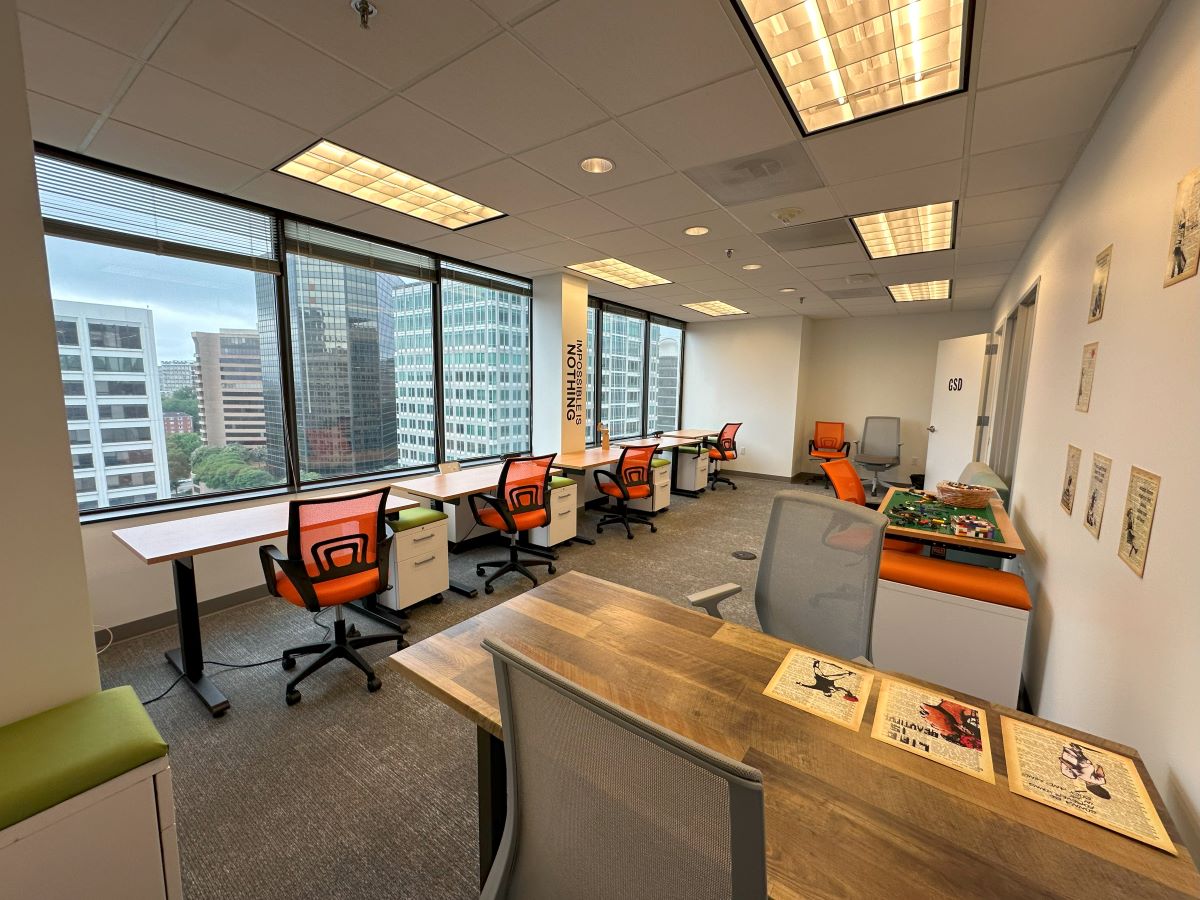 A row of desks with orange chairs sits in front of a window. Tables line the opposing wall. 