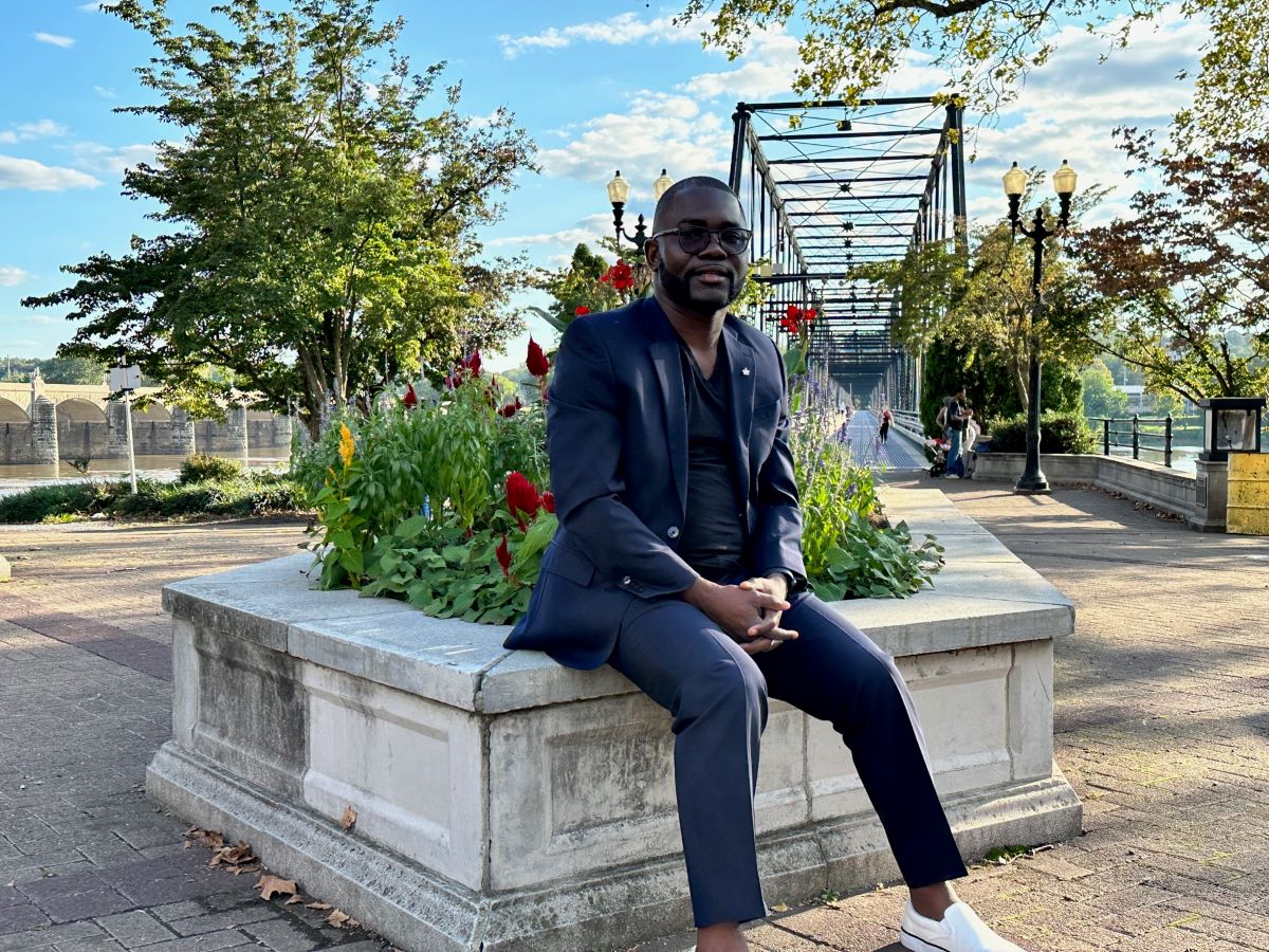 Michael Akinwumi sits on a stone garden wall on a pathway in front of a bridge. He wears a blue suit. 