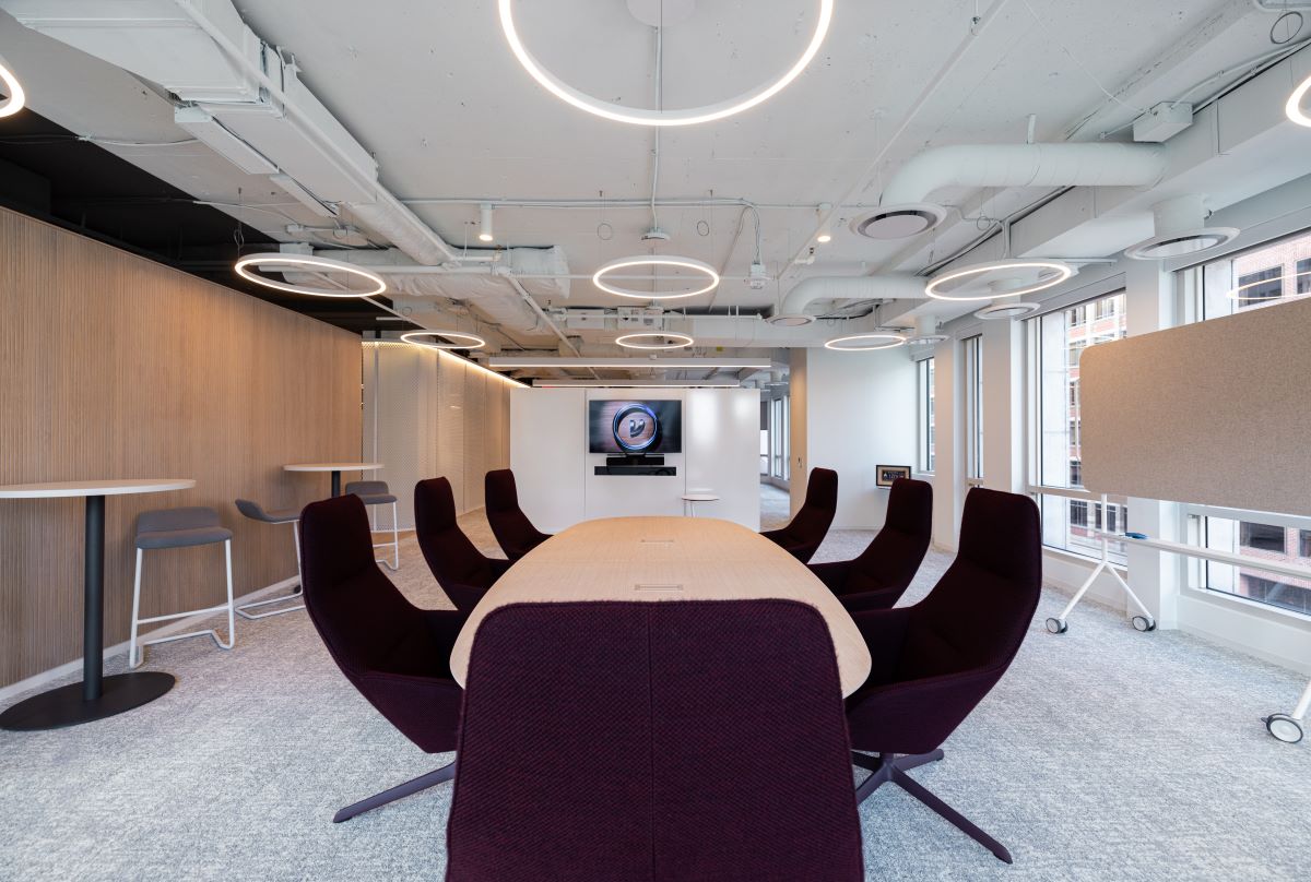 A conference room with dark chairs, white flooring and light wood under ring lights.