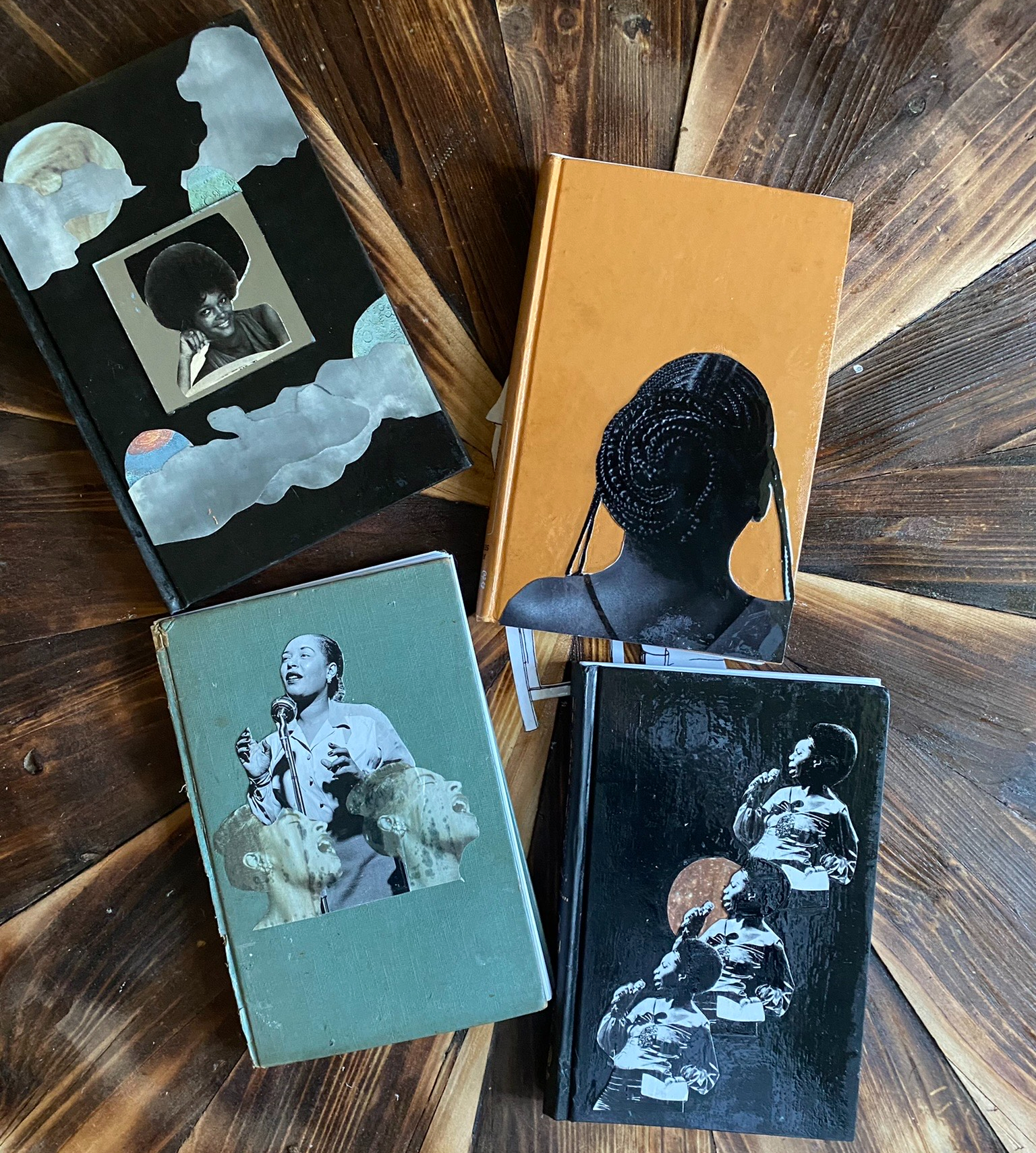 Four journals sit on a wood table. In the upper left corner, a black journal features a collage of clouds, the moon, and a Black woman. In the upper right corner, a yellow journal features an image of the back of a Black woman's head, with her hair braided into small braids and those braids twisted into a bun. In the lower right corner, a black journal features a collage of a woman singing. In the lower left corner, a green journal features a collage of another woman singing.