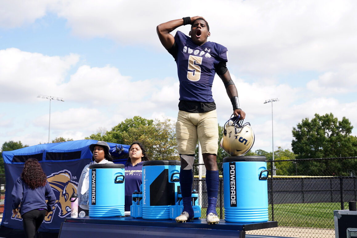 A football player stands in front of coolers holding his helmet. His hand is on his head and his expression is shocked.