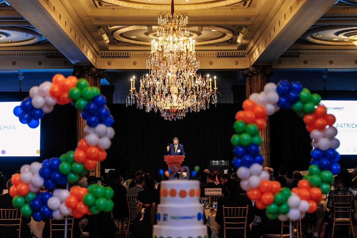 A man speaks onstage behind balloons arranged to spell the number 20.